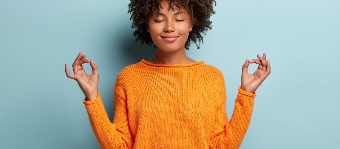 Mindful peaceful Afro American woman meditates indoor, keeps hands in mudra gesture, has eyes closed, tries to relax after long hours of working, holds fingers in yoga sign, isolated on blue wall