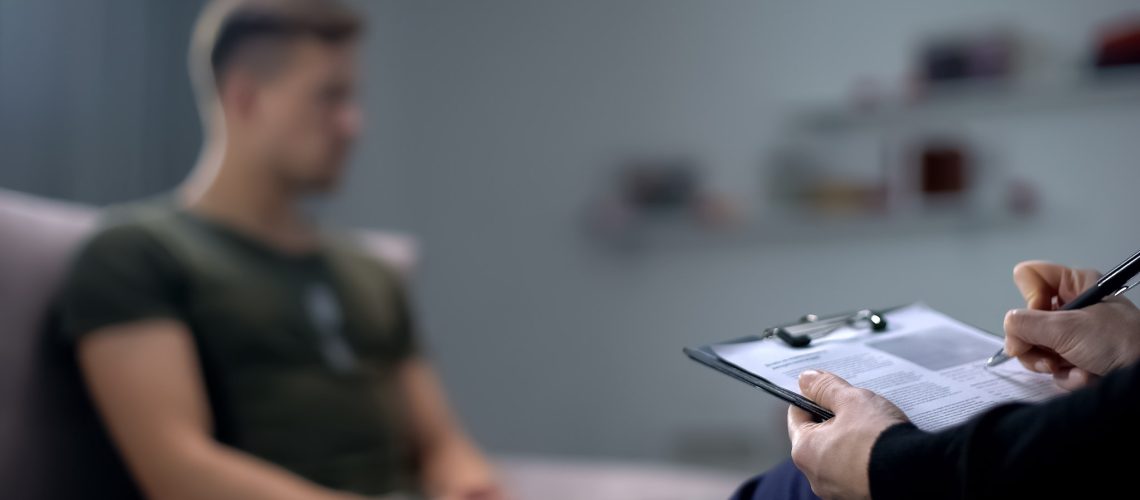 Psychologist making notes during therapy session with sad male soldier, PTSD